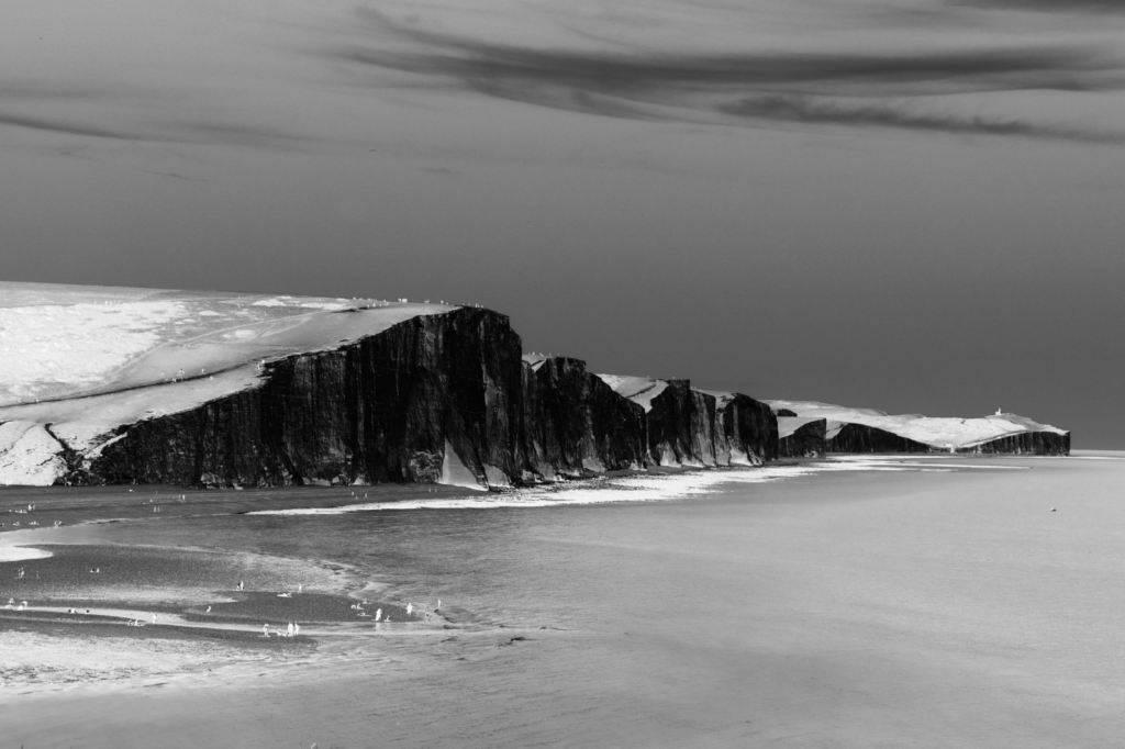 Black and white photo of tall, steep cliffs by a calm shoreline, with waves meeting the sandy beach and cloudy sky overhead.