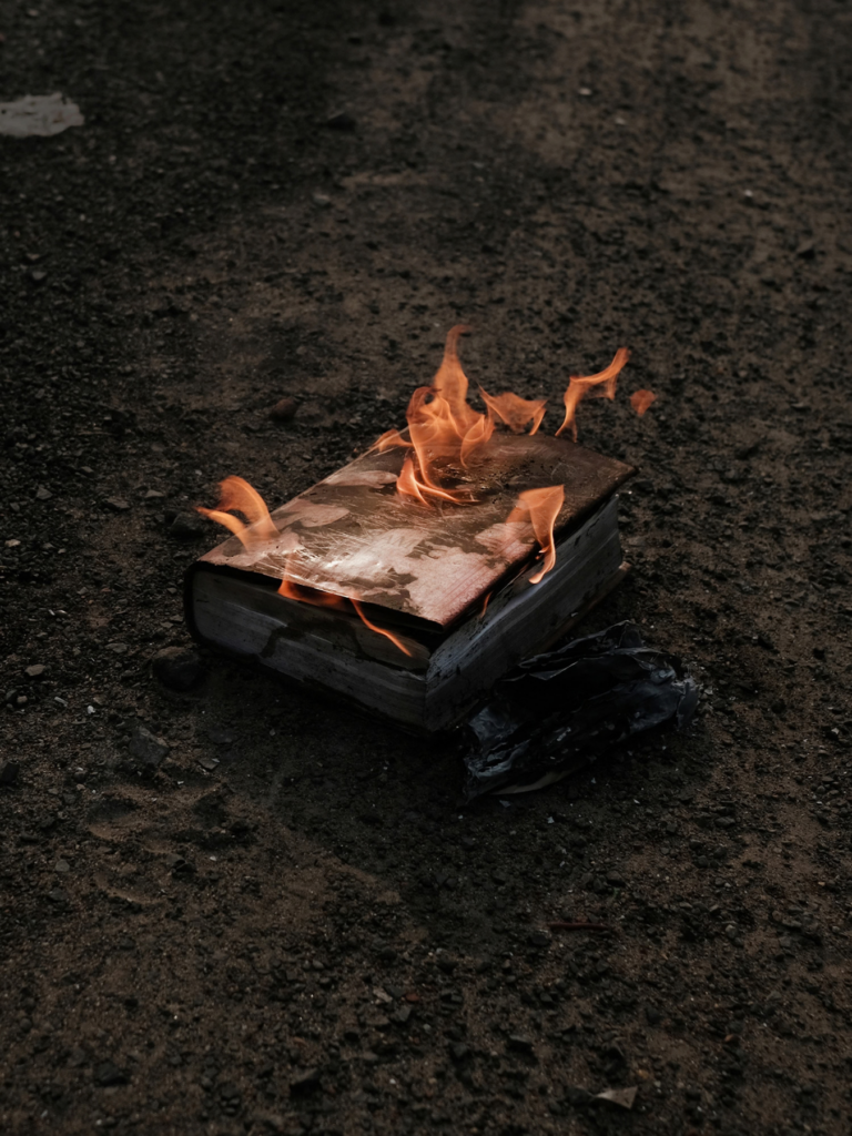 A photograph of a thick book burning on dark, rough ground, with orange flames curling above its charred pages.
