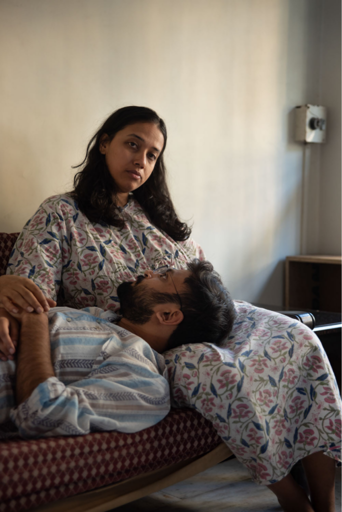 A woman sits on a patterned sofa staring directly to camera, a man is laying across her lap looking up at her