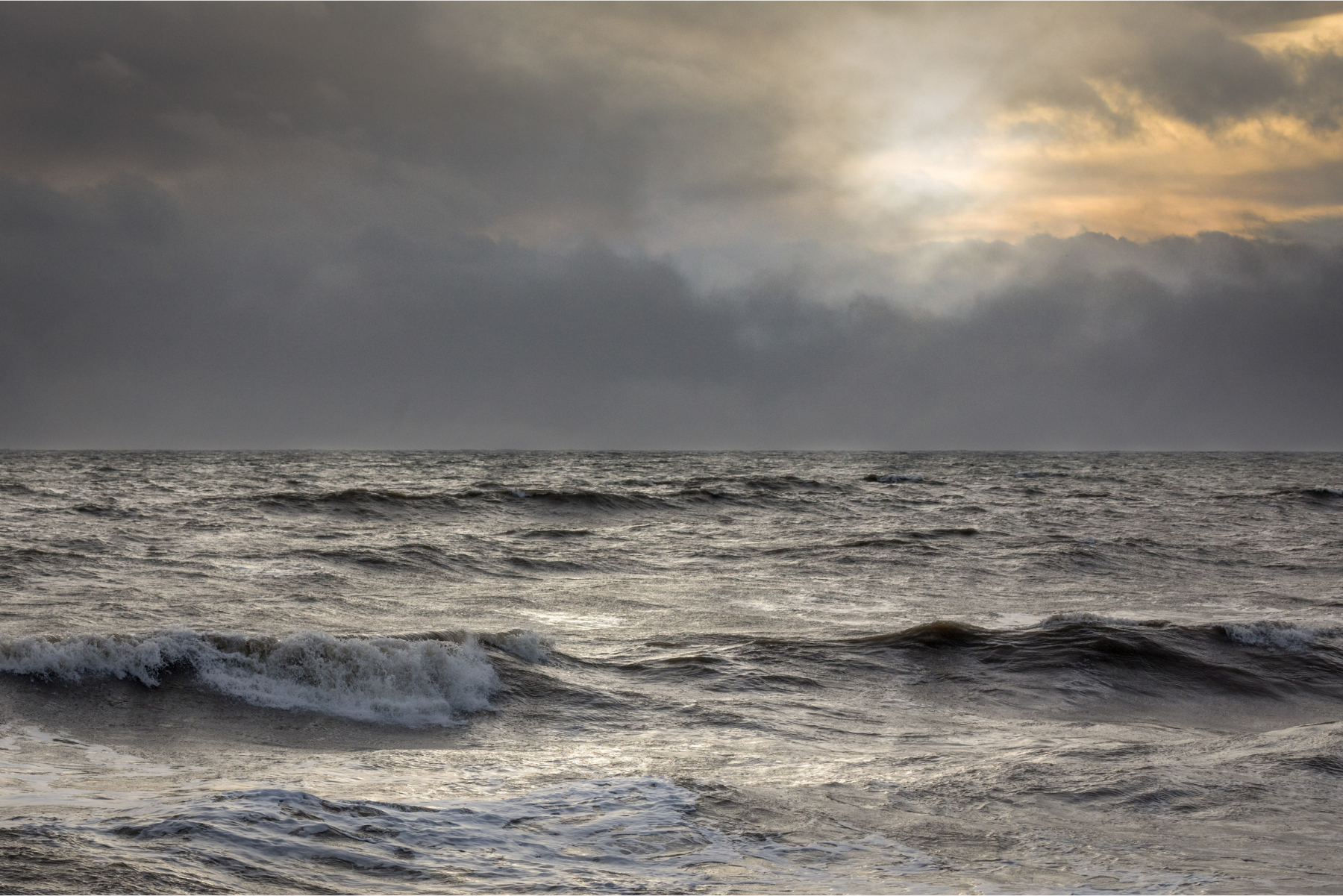 A photograph. Choppy ocean waves roll beneath dark storm clouds, with sunlight breaking through near the horizon.