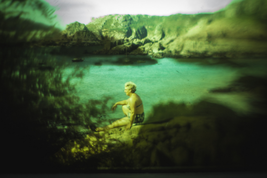 A person sits at the edge of a clear turquoise alpine lake, surrounded by green mountains and shadowed foliage;