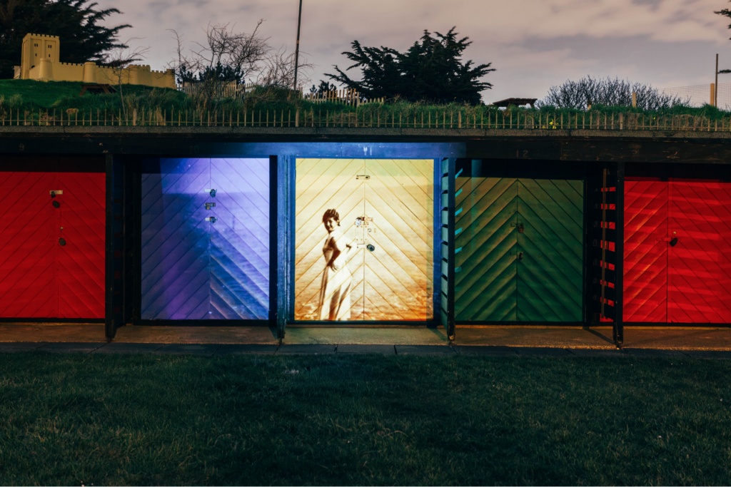 A row of colorful beach huts at night, with a vintage photograph of a woman projected onto the central door, set against a grassy hill and trees.